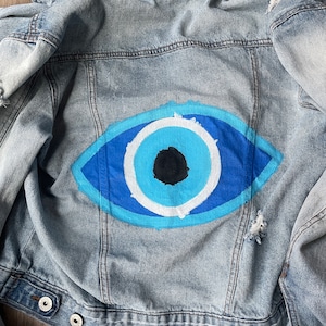 May include: Light blue denim jacket with a painted blue eye design on the back. The eye features shades of blue, white, and a black pupil. The jacket has a distressed look with ripped details.