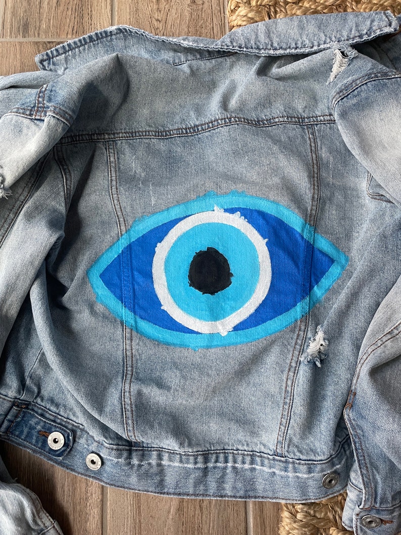 May include: Light blue denim jacket with a painted blue eye design on the back. The eye features shades of blue, white, and a black pupil. The jacket has a distressed look with ripped details.