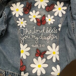 May include: A denim jacket with a hand-painted design featuring white daisies, red butterflies, and the text "The Lord bless you, my daughter - Ruth 3:10" in white script. The jacket is a light blue wash.