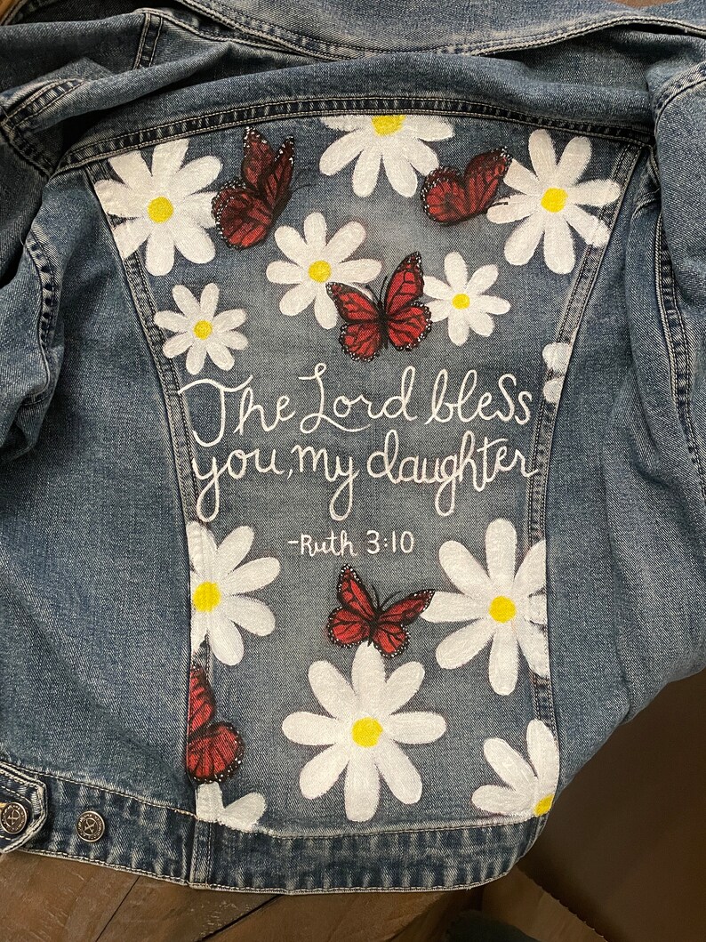 May include: A denim jacket with a hand-painted design featuring white daisies, red butterflies, and the text "The Lord bless you, my daughter - Ruth 3:10" in white script. The jacket is a light blue wash.