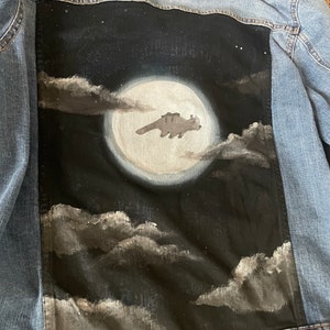 May include: A light blue denim jacket with a hand-painted design on the back. The artwork depicts a full moon, clouds, and a creature silhouette. The design is painted in shades of black, gray, and white.