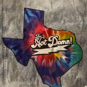 May include: Light blue denim jacket with a colorful tie-dye Texas-shaped patch. The patch features the text "The Hot Damn!" in white script. The jacket has a classic design with button closures and pockets.