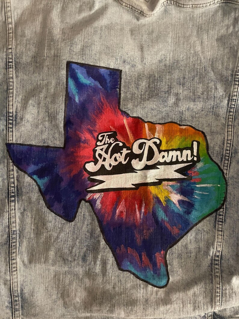 May include: Light blue denim jacket with a colorful tie-dye Texas-shaped patch. The patch features the text "The Hot Damn!" in white script. The jacket has a classic design with button closures and pockets.