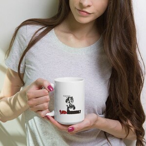 May include: White ceramic mug featuring a black and red graphic of a figure with a weapon and the text "FUMBLE THOROUGHLY". The mug has a large handle and a glossy finish. The mug is held by a person.