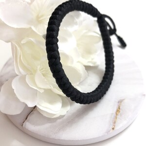 May include: A black braided bracelet with a sliding knot closure. The bracelet is made of a thick, woven material and is displayed on a white and marble-patterned surface, with white flowers in the background.