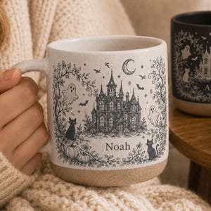 May include: A cream-colored ceramic mug featuring a black ink illustration of a haunted house, ghosts, cats, pumpkins, and a crescent moon, with the name Noah printed below the house. A second black mug with a similar design and the name Amelia is visible.