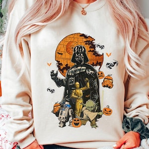 May include: Cream-colored sweatshirt featuring a Halloween-themed Star Wars graphic. The design includes Darth Vader, other characters, and a Death Star with pumpkins and bats. A festive and unique apparel item.