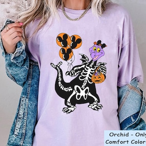May include: An orchid-coloured t-shirt featuring a graphic of a purple dragon wearing a skeleton costume, holding three orange Mickey Mouse-shaped balloons and an orange jack-o'-lantern bucket. The shirt is layered under a blue denim jacket.