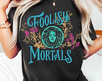May include: A black t-shirt featuring the text FOOLISH MORTALS in a light blue script font. The design includes a central circular graphic of a face in light blue, surrounded by gold swirling vines and pink rose illustrations.