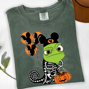 May include: A dark olive green t-shirt featuring a graphic of a green chameleon wearing a black skeleton costume and a black hat with an orange mouse-ear icon. The chameleon holds three orange balloons with black mouse-ear silhouettes and a pumpkin bucket filled with sweets.