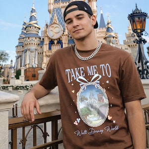 May include: Brown t-shirt with the text "TAKE ME TO" above a framed image of a castle and the words "Walt Disney World". The shirt has a crew neck and short sleeves. The person is wearing a black baseball cap and a silver chain.