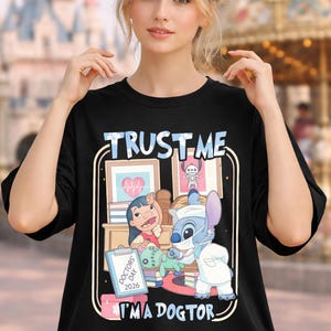 Pu&ograve; includere: T-shirt nero con grafica a cartoni animati di Lilo e Stitch a tema medico. La maglietta presenta la scritta "TRUST ME I'M A DOCTOR" in bianco e blu. Il design include illustrazioni mediche e la scritta "DOCTORS DAY 2026".