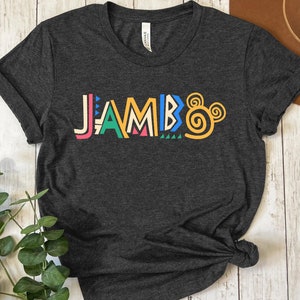 Puede incluir: Una camiseta gris oscuro con la palabra "JAMBO" impresa en letras estilizadas de colores. Las letras est&aacute;n delineadas en negro y tienen un patr&oacute;n geom&eacute;trico. La palabra est&aacute; centrada en la camiseta y est&aacute; ligeramente inclinada hacia la derecha.