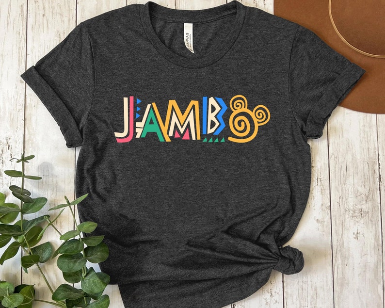 Puede incluir: Una camiseta gris oscuro con la palabra "JAMBO" impresa en letras estilizadas de colores. Las letras est&aacute;n delineadas en negro y tienen un patr&oacute;n geom&eacute;trico. La palabra est&aacute; centrada en la camiseta y est&aacute; ligeramente inclinada hacia la derecha.