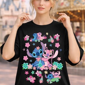 May include: Black t-shirt featuring a colorful graphic of the Disney characters Stitch and Angel surrounded by flowers. The design includes Stitch and Angel in various poses, with pink and blue floral accents.