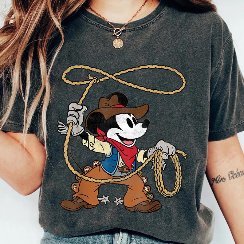 May include: Dark gray t-shirt featuring a cartoon cowboy Mickey Mouse holding a lasso. Mickey is wearing a brown cowboy hat, vest, and pants, with a red bandana. The design includes two white star accents.