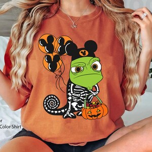 May include: A yam-coloured t-shirt featuring a graphic of a green chameleon wearing a black mouse-ear hat, holding orange balloons with mouse-ear silhouettes, and carrying a jack-o'-lantern bucket filled with colourful candy.