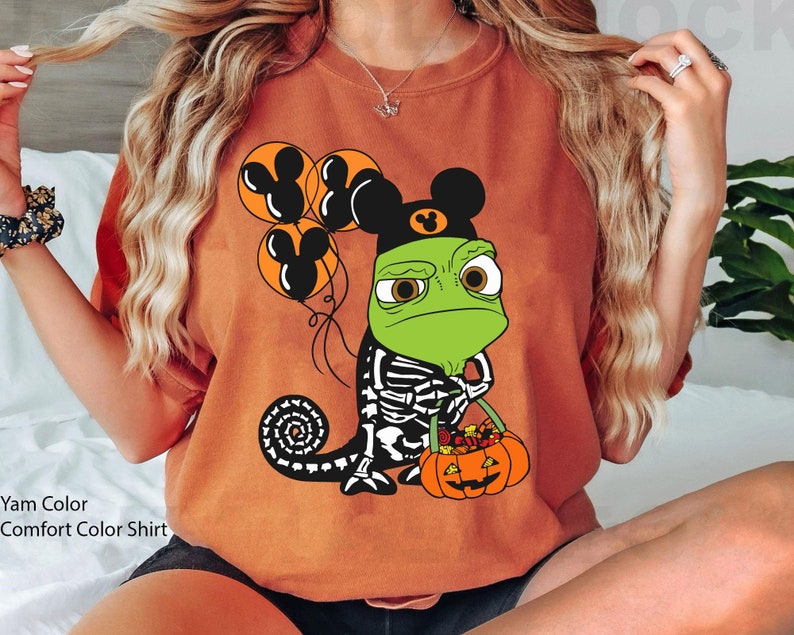 May include: A yam-coloured t-shirt featuring a graphic of a green chameleon wearing a black mouse-ear hat, holding orange balloons with mouse-ear silhouettes, and carrying a jack-o'-lantern bucket filled with colourful candy.