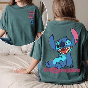 May include: Teal-colored t-shirt featuring a cartoon Stitch design. The front has a pink Mickey Mouse head with Stitch. The back displays a large Stitch graphic with the text "Walt Disney World" in pink. A comfortable, casual tee.