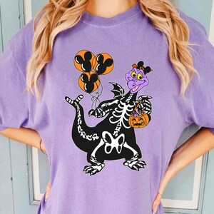 May include: A light purple t-shirt featuring a graphic of a purple dragon wearing a black skeleton costume. The dragon holds three orange balloons with black Mickey Mouse silhouettes and carries an orange jack-o'-lantern candy bucket.