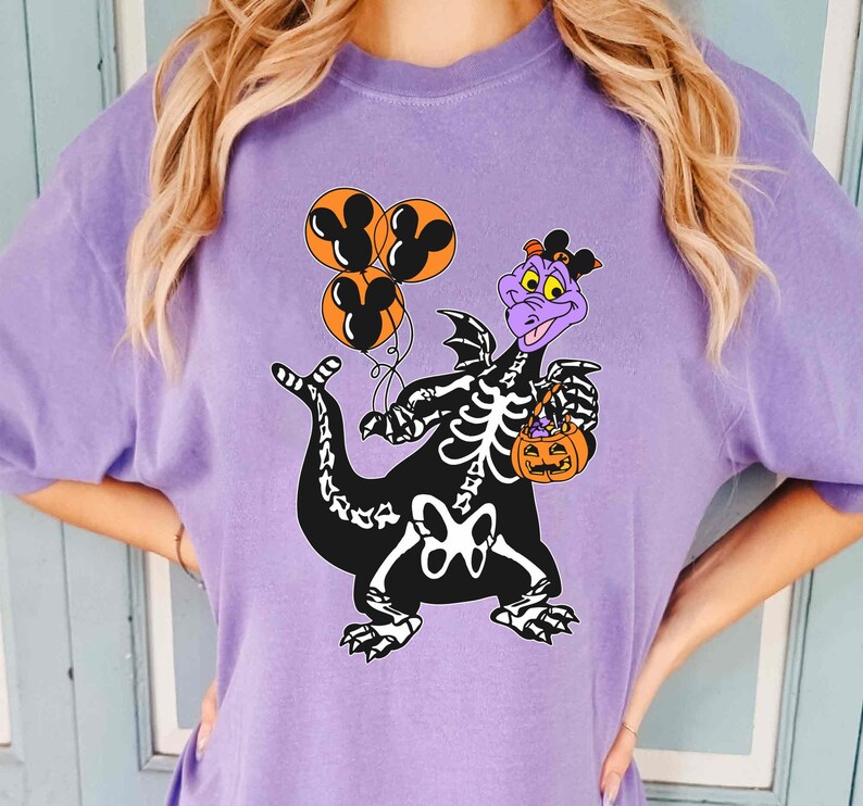 May include: A light purple t-shirt featuring a graphic of a purple dragon wearing a black skeleton costume. The dragon holds three orange balloons with black Mickey Mouse silhouettes and carries an orange jack-o'-lantern candy bucket.
