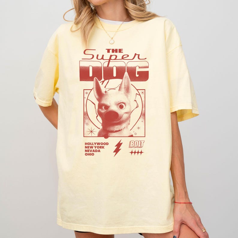 May include: A light yellow t-shirt featuring a graphic of a dog with the text "THE SUPER DOG" in red. Additional text includes "HOLLYWOOD NEW YORK NEVADA OHIO" and "BOLT".