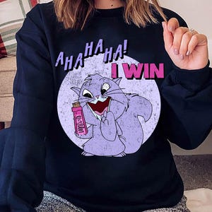 Puede incluir: Una sudadera azul oscuro con un gato de dibujos animados morado que sostiene una botella rosa. El texto "AHA HA HA! I WIN" está impreso en la sudadera.