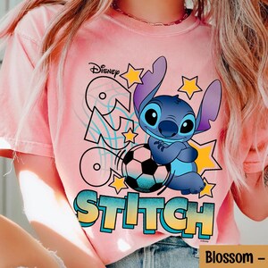 May include: Pink t-shirt featuring a graphic of Stitch from Disney's Lilo & Stitch. Stitch is depicted in blue, holding a soccer ball, with the word "Stitch" in a colorful font. The Disney logo is visible at the top.