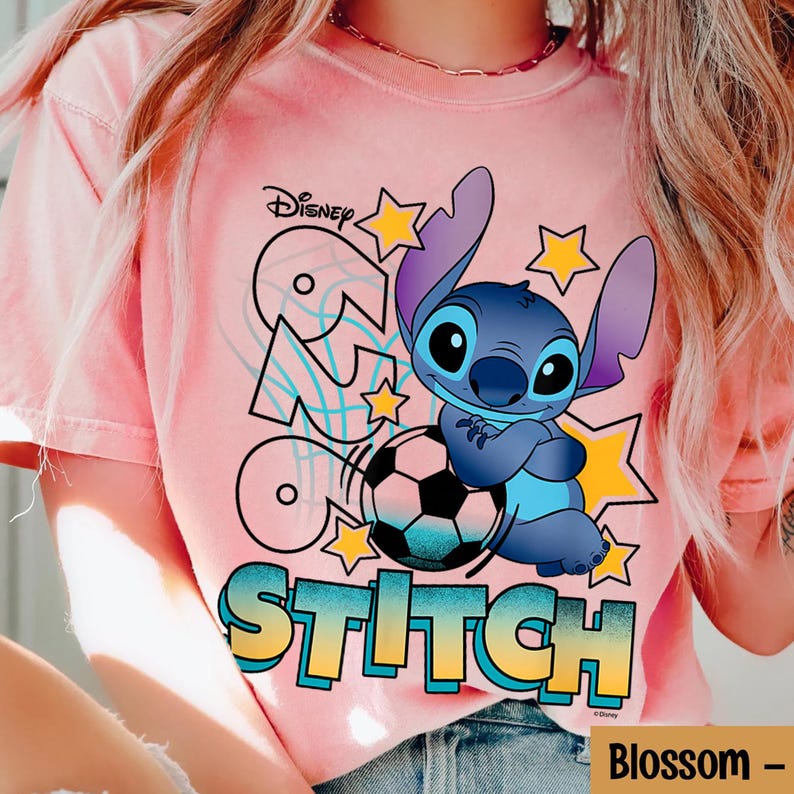 May include: Pink t-shirt featuring a graphic of Stitch from Disney's Lilo & Stitch. Stitch is depicted in blue, holding a soccer ball, with the word "Stitch" in a colorful font. The Disney logo is visible at the top.