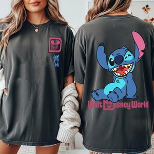 May include: Dark gray t-shirt featuring a cartoon Stitch character on the back, winking and smiling. The front has a pink "D" logo with a globe and a small Stitch graphic. The text "Walt Disney World" is below Stitch.