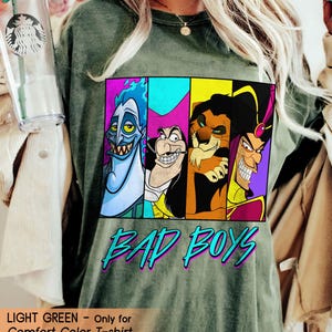 May include: Light green Comfort Color T-shirt featuring a graphic of four cartoon villains in colorful panels. The words "BAD BOYS" are printed in turquoise with a purple outline. The shirt is a relaxed fit.