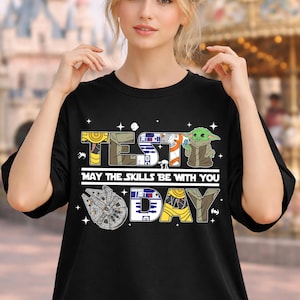 Puede incluir: Camiseta negra con un diseño temático de Star Wars que dice "TEST DAY". El diseño incluye imágenes de R2-D2, Grogu y el Halcón Milenario. La frase "MAY THE SKILLS BE WITH YOU" también está impresa en la camiseta.
