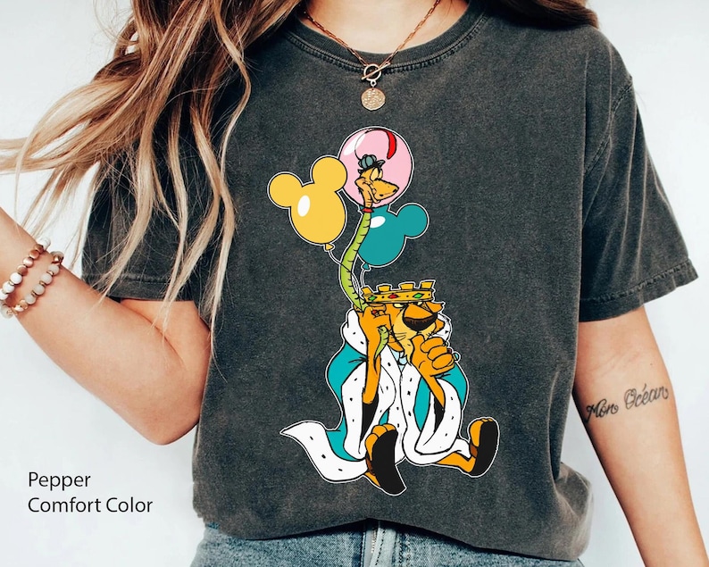 May include: A black t-shirt with a cartoon lion wearing a crown and holding balloons. The balloons are yellow, blue, and pink. The lion is wearing a blue and white robe. The text "More Ocean" is written on the right sleeve of the shirt.