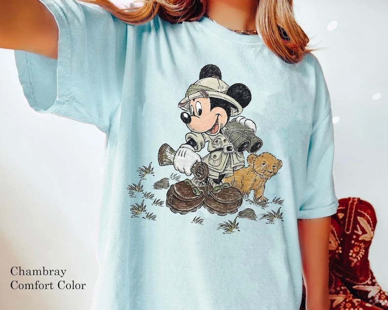 May include: A light blue t-shirt with a graphic of Mickey Mouse dressed as a safari explorer with a lion cub. The text "Chambray Comfort Color" is printed on the bottom left corner of the shirt.