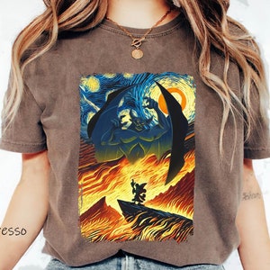 May include: A brown t-shirt with a graphic print of a scene from the Disney movie *The Hunchback of Notre Dame*. The scene depicts the villain, Frollo, in a fiery inferno, with the sky above resembling Vincent van Gogh's *Starry Night* painting.