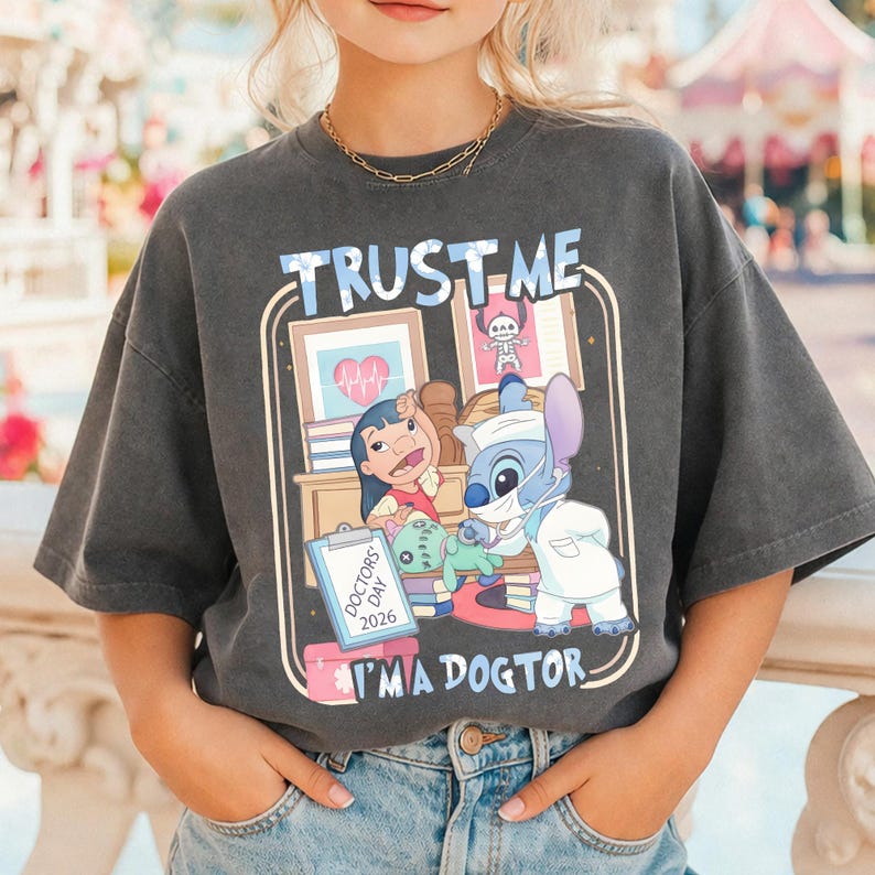 Pu&ograve; includere: T-shirt grigio scuro con grafica a cartoni animati. La grafica include il testo "TRUST ME" e "I'M A DOGTOR", insieme ai personaggi di Lilo e Stitch in una scena a tema medico. Include illustrazioni mediche e un cartello "DOCTOR'S DAY 2026".