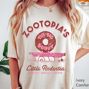 Puede incluir: Camiseta color marfil con un gr&aacute;fico de una rosquilla y el texto "Zootopia's The Big Donut" en marr&oacute;n y rosa. Debajo, un carrito de comida rosa y blanco con el texto "Little Rodentia" y "Donuts, Bagels, Coffee".