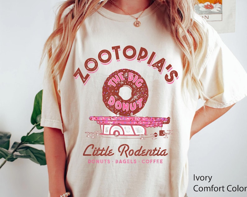 Puede incluir: Camiseta color marfil con un gr&aacute;fico de una rosquilla y el texto "Zootopia's The Big Donut" en marr&oacute;n y rosa. Debajo, un carrito de comida rosa y blanco con el texto "Little Rodentia" y "Donuts, Bagels, Coffee".