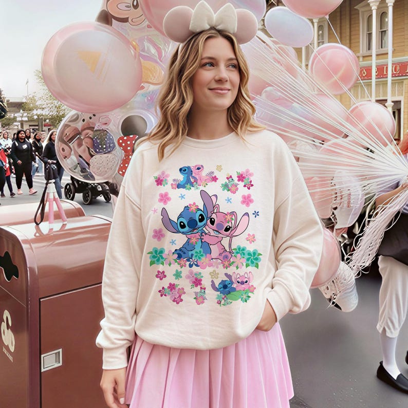 May include: Cream-colored sweatshirt featuring a colorful graphic of Stitch and Angel characters surrounded by flowers. The sweatshirt is paired with a pink pleated skirt. The background includes pink balloons and a theme park setting.