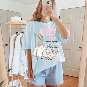 May include: A light blue t-shirt with a graphic of Bo Peep from Toy Story. The graphic includes Bo Peep holding a shepherd's crook and three sheep. The text on the shirt reads "Bo Peep & Sheep Porcelain Figure Set Only at Select Toy Stores!"