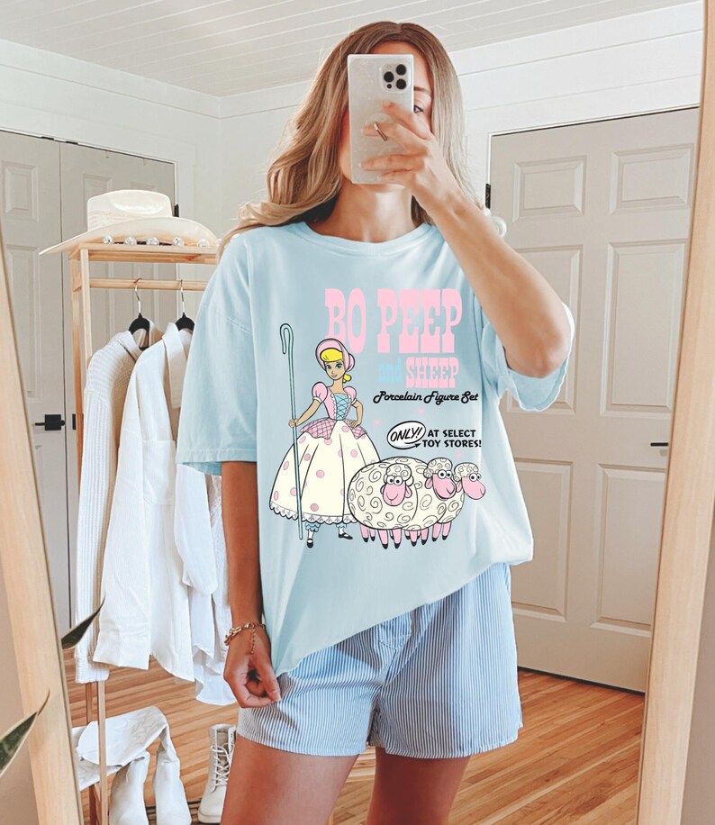 May include: A light blue t-shirt with a graphic of Bo Peep from Toy Story. The graphic includes Bo Peep holding a shepherd's crook and three sheep. The text on the shirt reads "Bo Peep & Sheep Porcelain Figure Set Only at Select Toy Stores!"