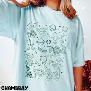 Puede incluir: Camiseta azul claro con un dise&ntilde;o en contorno verde de personajes de la pel&iacute;cula Toy Story. El dise&ntilde;o incluye a Woody, Buzz Lightyear, Rex y otros personajes. La palabra "CHAMBRAY" est&aacute; impresa en la parte inferior izquierda.