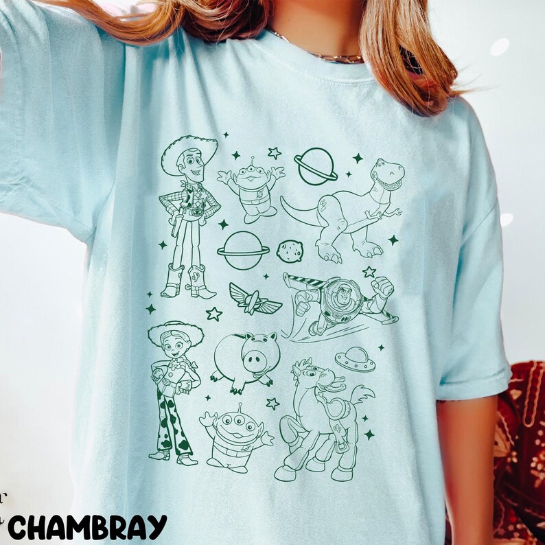 Puede incluir: Camiseta azul claro con un dise&ntilde;o en contorno verde de personajes de la pel&iacute;cula Toy Story. El dise&ntilde;o incluye a Woody, Buzz Lightyear, Rex y otros personajes. La palabra "CHAMBRAY" est&aacute; impresa en la parte inferior izquierda.