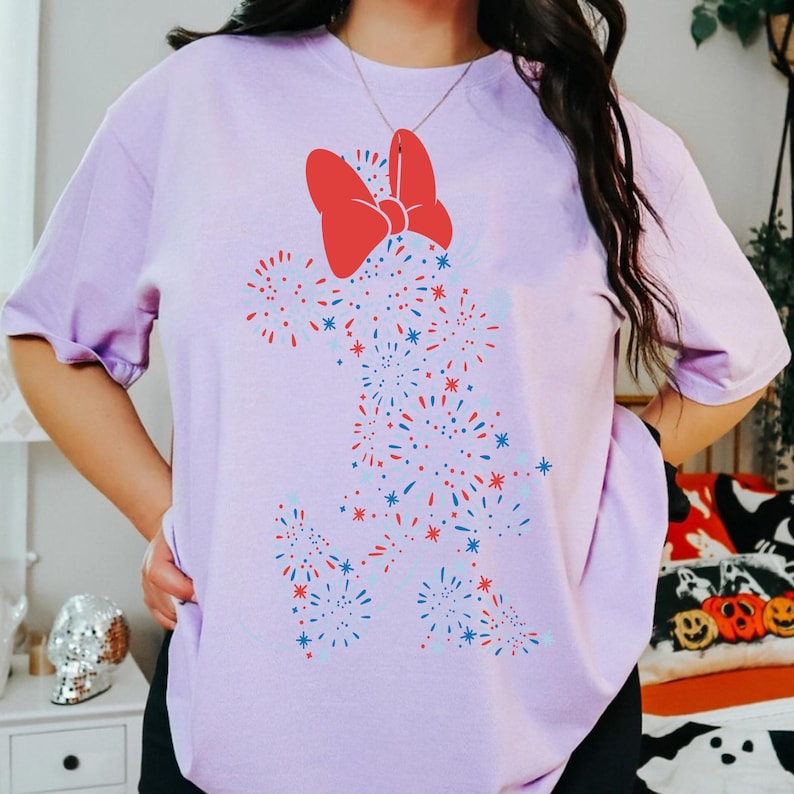 May include: A light purple t-shirt featuring a red bow and a firework design in red, white, and blue. The graphic is shaped like a character. The shirt has short sleeves and a relaxed fit.