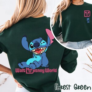 May include: Forest green sweatshirt featuring a smiling blue cartoon character with large ears and a wide grin. The back of the sweatshirt displays the character and the words "Walt Disney World". A smaller graphic is on the upper chest.