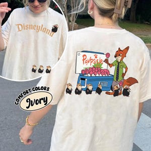 Puede incluir: Camiseta de color marfil con "Disneyland" impreso en marr&oacute;n. La camiseta presenta personajes de dibujos animados y el texto "Comfort Colors Ivory". La parte trasera de la camiseta tiene un zorro de dibujos animados vendiendo paletas.