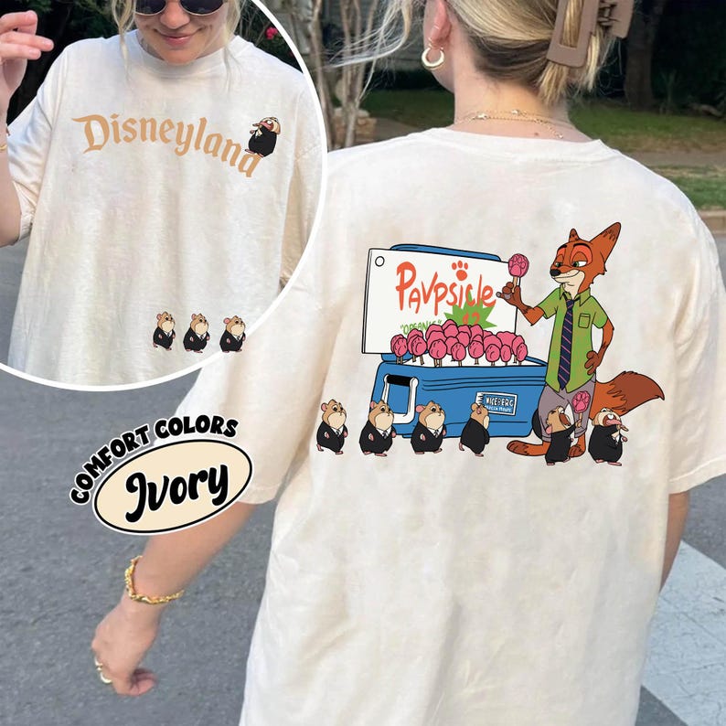 Puede incluir: Camiseta de color marfil con "Disneyland" impreso en marr&oacute;n. La camiseta presenta personajes de dibujos animados y el texto "Comfort Colors Ivory". La parte trasera de la camiseta tiene un zorro de dibujos animados vendiendo paletas.