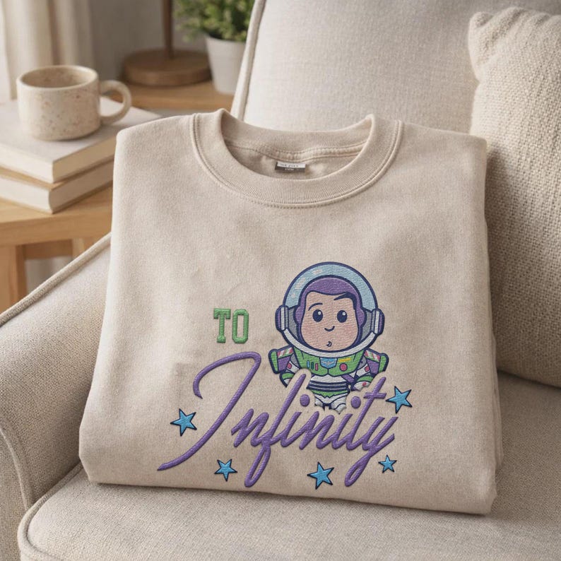 Puede incluir: Sudadera beige con una ilustraci&oacute;n de dibujos animados de un astronauta y el texto "TO Infinity" en escritura p&uacute;rpura, rodeado de estrellas azules. La sudadera doblada est&aacute; sobre un sill&oacute;n beige.