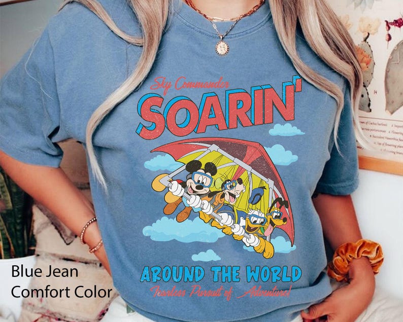 Puede incluir: Una camiseta azul vaquero con un gr&aacute;fico de Mickey Mouse y sus amigos volando en un ala delta. El texto "Soarin' Around the World" est&aacute; impreso en la camiseta.