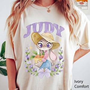 May include: Ivory t-shirt with a cartoon bunny character named Judy. The bunny wears a straw hat and holds a basket of carrots, surrounded by a floral wreath. The name "JUDY" is arched above the character in purple.